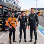 A realistic photograph of Formula 1 drivers Lando Norris (left, orange suit), Kimi Antonelli (middle, black suit, raised index finger), and George Russell (right, black suit, tallest) standing in the Zandvoort pit lane. Norris in McLaren gear, Antonelli and Russell in Mercedes gear. Background shows pit garages, a McLaren sign, tire stacks with 'ZANDVOORT F1 DUTCH GRAND PRIX' text, and distant sand dunes under bright daylight. Full race suits and detailed environment.