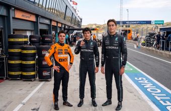 A realistic photograph of Formula 1 drivers Lando Norris (left, orange suit), Kimi Antonelli (middle, black suit, raised index finger), and George Russell (right, black suit, tallest) standing in the Zandvoort pit lane. Norris in McLaren gear, Antonelli and Russell in Mercedes gear. Background shows pit garages, a McLaren sign, tire stacks with 'ZANDVOORT F1 DUTCH GRAND PRIX' text, and distant sand dunes under bright daylight. Full race suits and detailed environment.