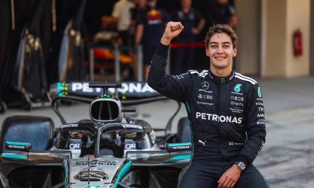 Formula 1 driver George Russell sitting on his Mercedes F1 car in the pit lane, smiling and raising his right fist in celebration of a first-place qualifying result.