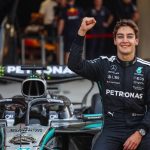 Formula 1 driver George Russell sitting on his Mercedes F1 car in the pit lane, smiling and raising his right fist in celebration of a first-place qualifying result.