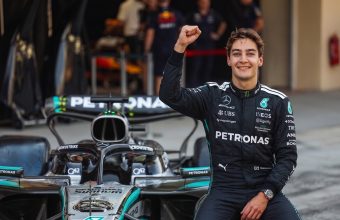 Formula 1 driver George Russell sitting on his Mercedes F1 car in the pit lane, smiling and raising his right fist in celebration of a first-place qualifying result.