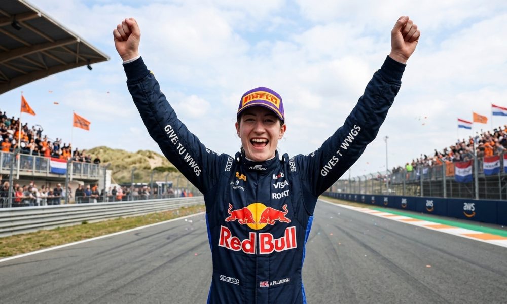Photorealistic image of a female Red Bull racing driver smiling joyfully and celebrating with both arms raised in victory at the Circuit Zandvoort Formula 1 track, wearing her blue racing suit and cap.