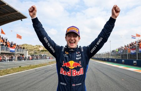 Photorealistic image of a female Red Bull racing driver smiling joyfully and celebrating with both arms raised in victory at the Circuit Zandvoort Formula 1 track, wearing her blue racing suit and cap.
