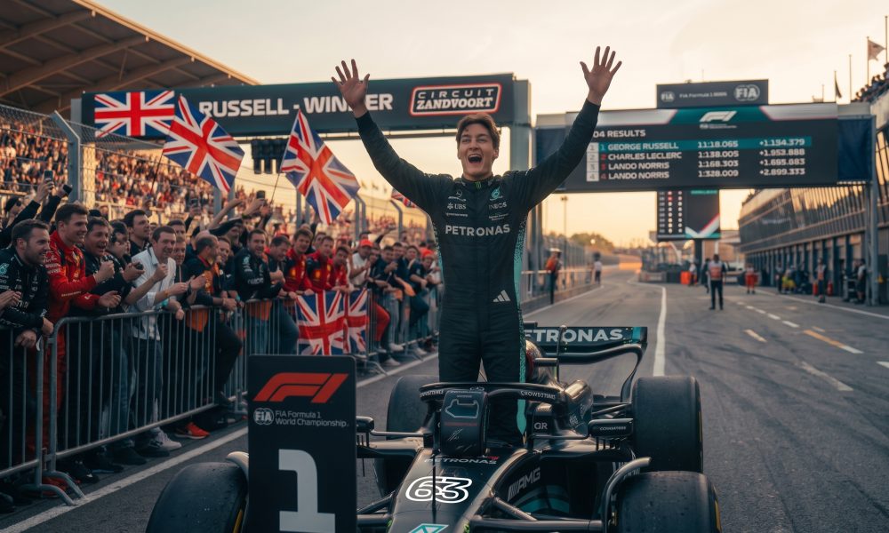 Photorealistic image of Formula 1 driver George Russell celebrating a victory on the Zandvoort circuit, standing with arms raised. Fans with Union Jack flags, Mercedes, and Ferrari apparel cheer behind him. A screen displays the podium: 1. George Russell, 2. Charles Leclerc, 3. Lando Norris.