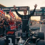 Photorealistic image of Formula 1 driver George Russell celebrating a victory on the Zandvoort circuit, standing with arms raised. Fans with Union Jack flags, Mercedes, and Ferrari apparel cheer behind him. A screen displays the podium: 1. George Russell, 2. Charles Leclerc, 3. Lando Norris.