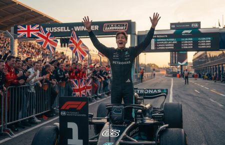 Photorealistic image of Formula 1 driver George Russell celebrating a victory on the Zandvoort circuit, standing with arms raised. Fans with Union Jack flags, Mercedes, and Ferrari apparel cheer behind him. A screen displays the podium: 1. George Russell, 2. Charles Leclerc, 3. Lando Norris.