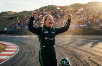 A blonde female Formula 1 driver in a black and green racing suit celebrating a victory at Circuit Zandvoort with her arms raised and a wide, joyful smile, with Dutch fans and flags visible in the background dunes at sunset.