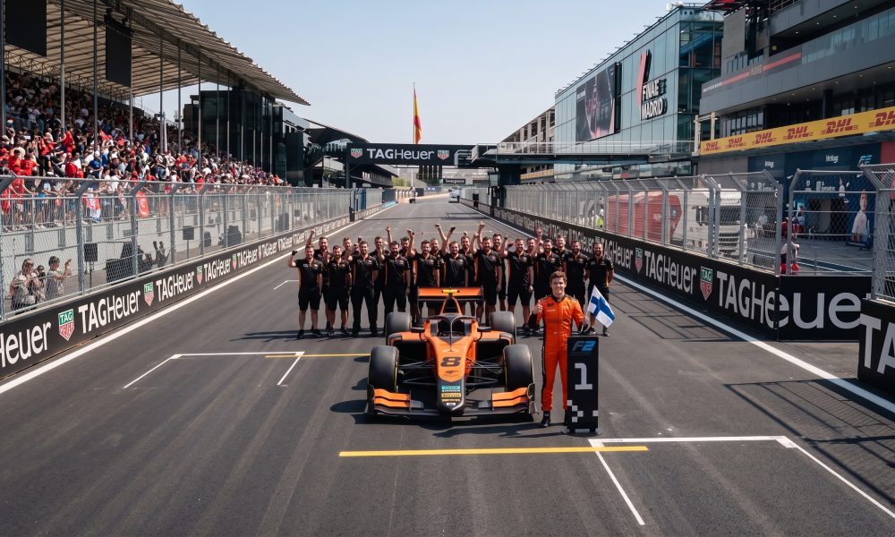 A smiling race car driver in an orange suit holds a Finnish flag next to his orange number 8 formula car on a race track starting grid. Behind the car, the entire pit crew is cheering, and fans are watching from the grandstands on the left.
