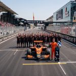 A smiling race car driver in an orange suit holds a Finnish flag next to his orange number 8 formula car on a race track starting grid. Behind the car, the entire pit crew is cheering, and fans are watching from the grandstands on the left.