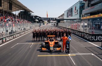 A smiling race car driver in an orange suit holds a Finnish flag next to his orange number 8 formula car on a race track starting grid. Behind the car, the entire pit crew is cheering, and fans are watching from the grandstands on the left.