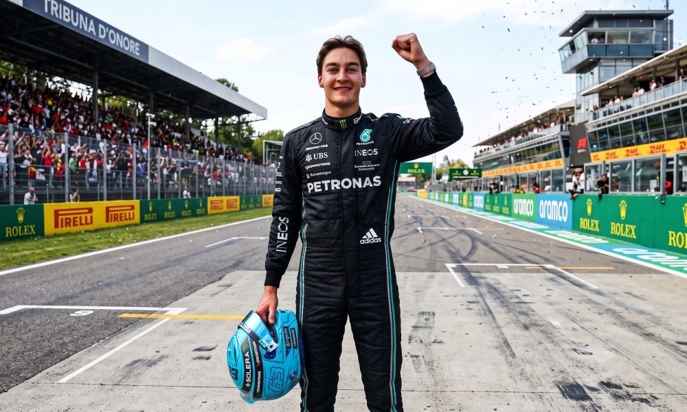 Formula 1 driver George Russell standing on the Monza circuit pit straight, raising his right fist in celebration. He is wearing a black Mercedes-AMG Petronas race suit and holding a light blue racing helmet in his left hand.