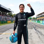 Formula 1 driver George Russell standing on the Monza circuit pit straight, raising his right fist in celebration. He is wearing a black Mercedes-AMG Petronas race suit and holding a light blue racing helmet in his left hand.