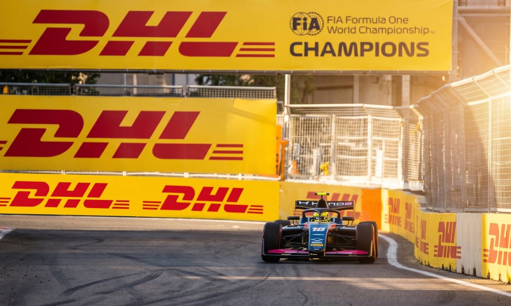 A blue and pink open-wheel race car, number 18, approaches a turn on a street circuit, flanked by massive yellow DHL and FIA Formula One Champions branded barriers under golden sunlight.
