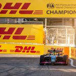 A blue and pink open-wheel race car, number 18, approaches a turn on a street circuit, flanked by massive yellow DHL and FIA Formula One Champions branded barriers under golden sunlight.