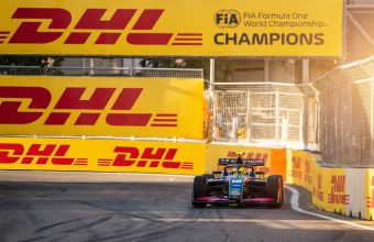 A blue and pink open-wheel race car, number 18, approaches a turn on a street circuit, flanked by massive yellow DHL and FIA Formula One Champions branded barriers under golden sunlight.