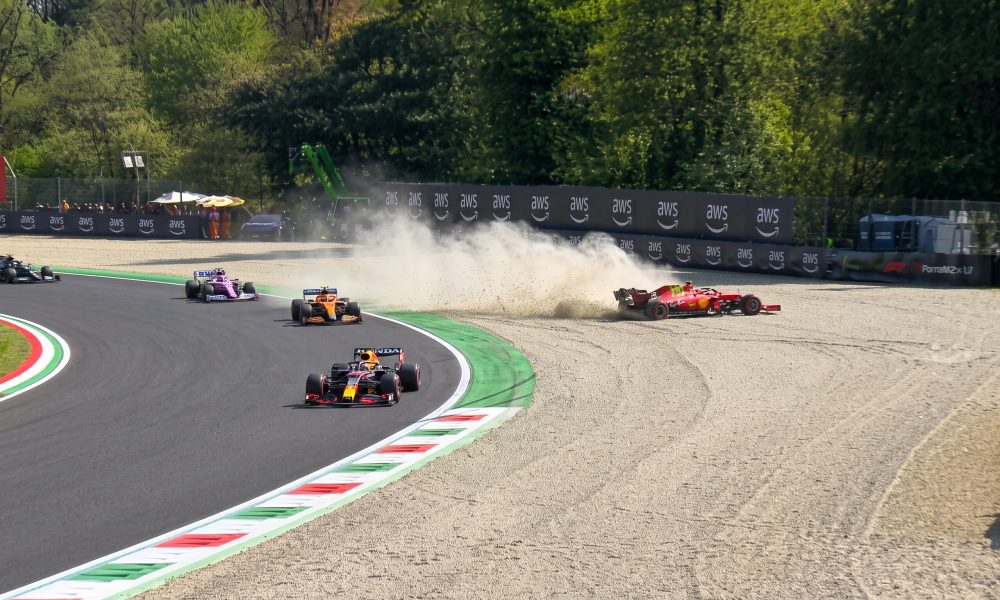 Action photograph of a Formula 1 race incident: a red Scuderia Ferrari car mired in a gravel trap, kicking up a large cloud of dust, while other F1 cars (Red Bull Racing, McLaren, Racing Point) continue navigating the corner on the asphalt track.
