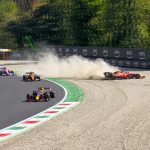 Action photograph of a Formula 1 race incident: a red Scuderia Ferrari car mired in a gravel trap, kicking up a large cloud of dust, while other F1 cars (Red Bull Racing, McLaren, Racing Point) continue navigating the corner on the asphalt track.