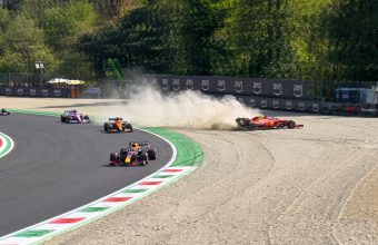 Action photograph of a Formula 1 race incident: a red Scuderia Ferrari car mired in a gravel trap, kicking up a large cloud of dust, while other F1 cars (Red Bull Racing, McLaren, Racing Point) continue navigating the corner on the asphalt track.