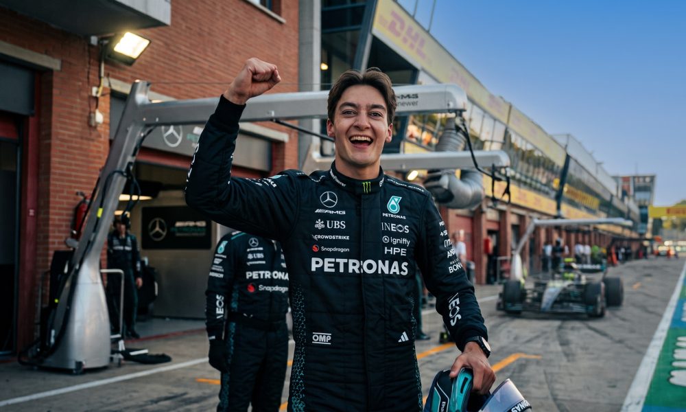A photographic medium shot of Formula 1 driver George Russell, wearing his black Mercedes-AMG Petronas team race suit, smiling broadly with a raised right fist in celebration. He is holding a helmet and standing in the pit lane. Legible logos include Mercedes-Benz, PETRONAS, UBS, INEOS, and CrowdStrike. The background is a racetrack garage area with other team members, out of focus.