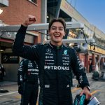 A photographic medium shot of Formula 1 driver George Russell, wearing his black Mercedes-AMG Petronas team race suit, smiling broadly with a raised right fist in celebration. He is holding a helmet and standing in the pit lane. Legible logos include Mercedes-Benz, PETRONAS, UBS, INEOS, and CrowdStrike. The background is a racetrack garage area with other team members, out of focus.