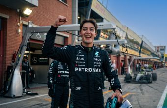 A photographic medium shot of Formula 1 driver George Russell, wearing his black Mercedes-AMG Petronas team race suit, smiling broadly with a raised right fist in celebration. He is holding a helmet and standing in the pit lane. Legible logos include Mercedes-Benz, PETRONAS, UBS, INEOS, and CrowdStrike. The background is a racetrack garage area with other team members, out of focus.