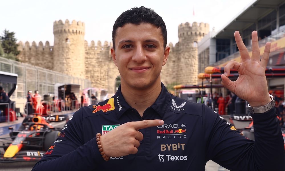 A smiling Oracle Red Bull Racing team member in uniform making hand gestures in the F1 paddock at Baku, with old city walls and race cars in the background.