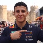 A smiling Oracle Red Bull Racing team member in uniform making hand gestures in the F1 paddock at Baku, with old city walls and race cars in the background.
