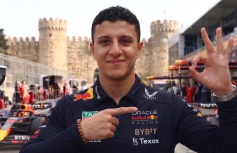 A smiling Oracle Red Bull Racing team member in uniform making hand gestures in the F1 paddock at Baku, with old city walls and race cars in the background.