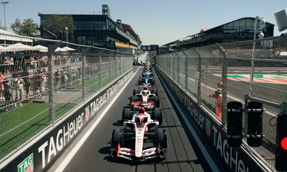 Formula One cars lined up on a street circuit starting grid, viewed from a low angle between tall metal catch fences with repeated 'TAG Heuer' branding. Large crowds of spectators watch from behind the fences on both sides. A modern trackside building with curved features is in the background under a clear blue sky. A vertical race start light with a bright red lamp is in the foreground to the right. The leading F1 car, number 31, is in focus. High-resolution photo.