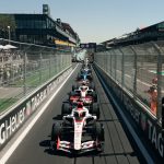 Formula One cars lined up on a street circuit starting grid, viewed from a low angle between tall metal catch fences with repeated 'TAG Heuer' branding. Large crowds of spectators watch from behind the fences on both sides. A modern trackside building with curved features is in the background under a clear blue sky. A vertical race start light with a bright red lamp is in the foreground to the right. The leading F1 car, number 31, is in focus. High-resolution photo.