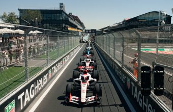 Formula One cars lined up on a street circuit starting grid, viewed from a low angle between tall metal catch fences with repeated 'TAG Heuer' branding. Large crowds of spectators watch from behind the fences on both sides. A modern trackside building with curved features is in the background under a clear blue sky. A vertical race start light with a bright red lamp is in the foreground to the right. The leading F1 car, number 31, is in focus. High-resolution photo.