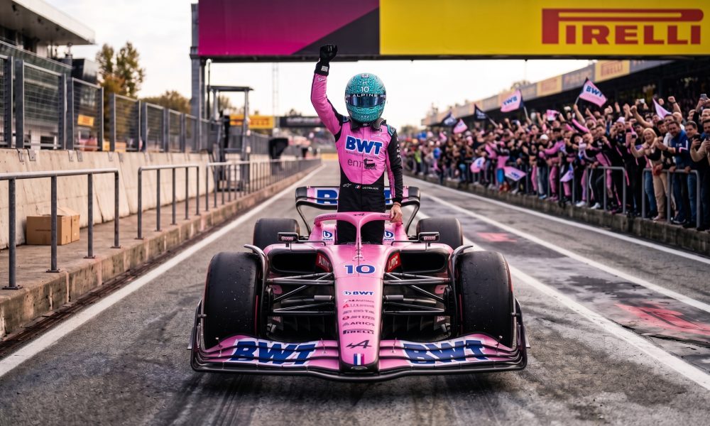 Formula 1 driver in a pink racing suit and blue helmet celebrating inside a pink BWT race car on the pit lane, with cheering fans in the background.