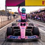 Formula 1 driver in a pink racing suit and blue helmet celebrating inside a pink BWT race car on the pit lane, with cheering fans in the background.