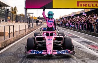 Formula 1 driver in a pink racing suit and blue helmet celebrating inside a pink BWT race car on the pit lane, with cheering fans in the background.