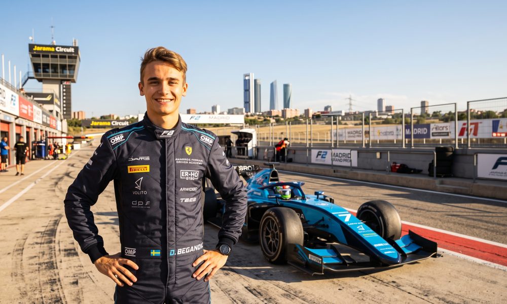 A wide-angle photorealistic landscape portrait of F2 driver Dino Beganovic in a dark blue race suit, smiling. He stands on the Jarama Circuit track, near Madrid. His light blue and black F2 car, number 7, is behind him. The distant Madrid skyline is visible under a warm, sunny sky. The suit and car show many detailed, legible sponsor logos.