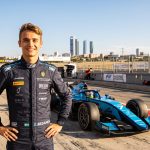 A wide-angle photorealistic landscape portrait of F2 driver Dino Beganovic in a dark blue race suit, smiling. He stands on the Jarama Circuit track, near Madrid. His light blue and black F2 car, number 7, is behind him. The distant Madrid skyline is visible under a warm, sunny sky. The suit and car show many detailed, legible sponsor logos.