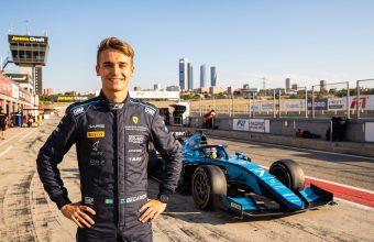 A wide-angle photorealistic landscape portrait of F2 driver Dino Beganovic in a dark blue race suit, smiling. He stands on the Jarama Circuit track, near Madrid. His light blue and black F2 car, number 7, is behind him. The distant Madrid skyline is visible under a warm, sunny sky. The suit and car show many detailed, legible sponsor logos.