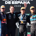 Four men on a Formula 1 podium, standing before a backdrop that reads 'GRAND PREMIO DE ESPAÑA' and '2026'. From left, a driver in a dark blue Red Bull suit waving; a team member with glasses in a black Mercedes polo; a driver in a black Mercedes suit with a gold medal and his arm raised high; and a driver in a dark grey Alpine suit waving. All three drivers wear Pirelli-branded caps. The backdrop features numerous sponsor logos.