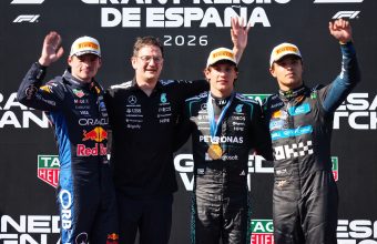Four men on a Formula 1 podium, standing before a backdrop that reads 'GRAND PREMIO DE ESPAÑA' and '2026'. From left, a driver in a dark blue Red Bull suit waving; a team member with glasses in a black Mercedes polo; a driver in a black Mercedes suit with a gold medal and his arm raised high; and a driver in a dark grey Alpine suit waving. All three drivers wear Pirelli-branded caps. The backdrop features numerous sponsor logos.